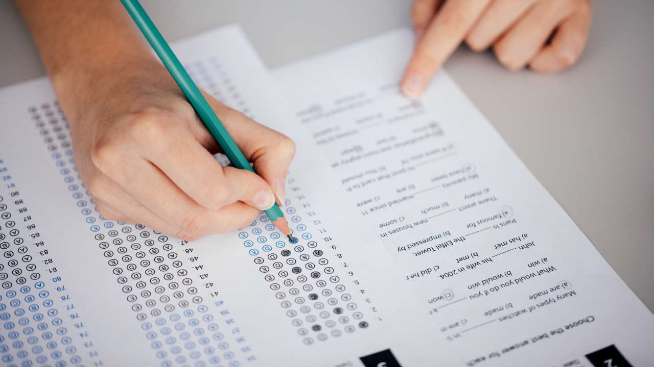 A person is filling in answers on a multiple-choice standardized test sheet with a pencil. Another hand, possibly belonging to one of the SAT English tutors, is pointing to a question on the test. The sheet has rows of bubbles and a column of questions visible.