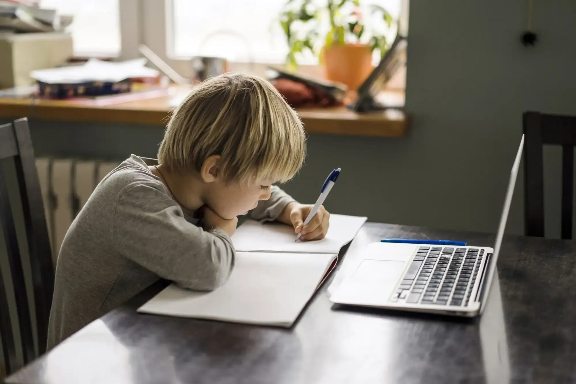 A child sits at a wooden table, writing in a notebook with a laptop nearby and a potted plant in the background, focused on their work.
