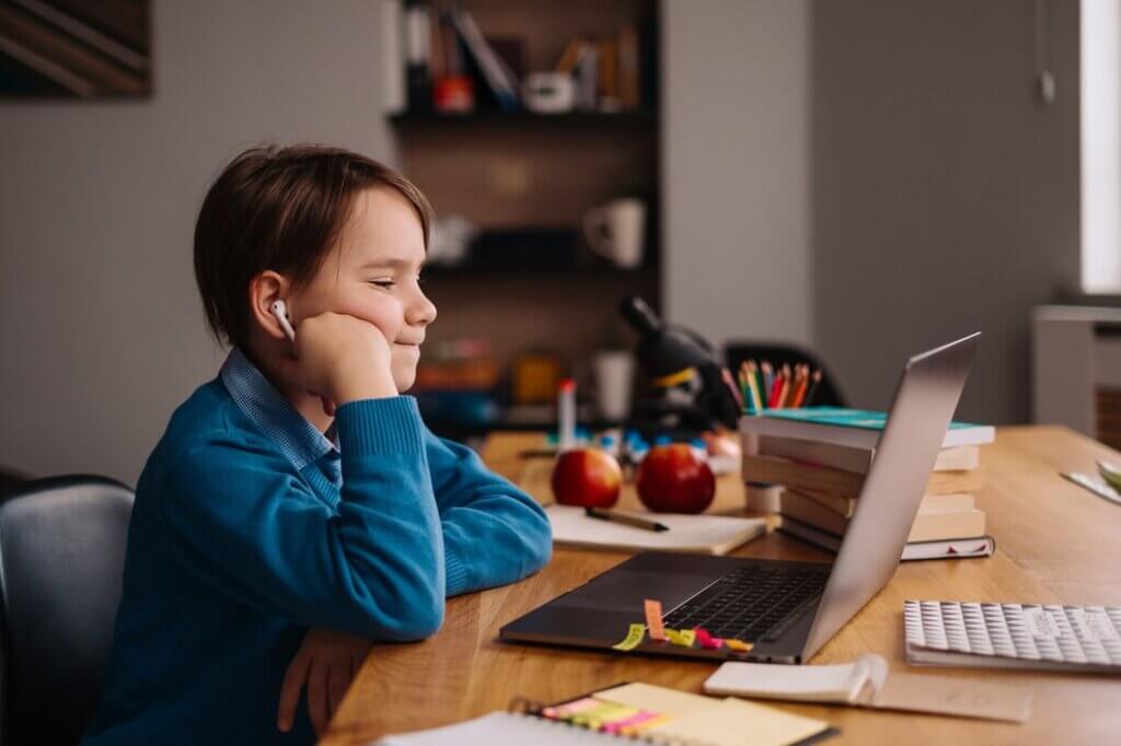 A child in a blue sweater sits at a desk, using a laptop surrounded by books, apples, and colorful stationery.