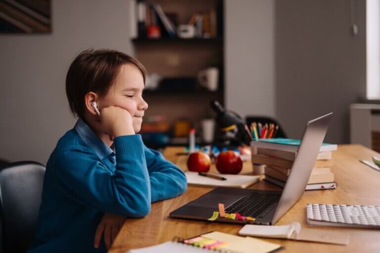 A child in a blue sweater sits at a desk, using a laptop surrounded by books, apples, and colorful stationery.