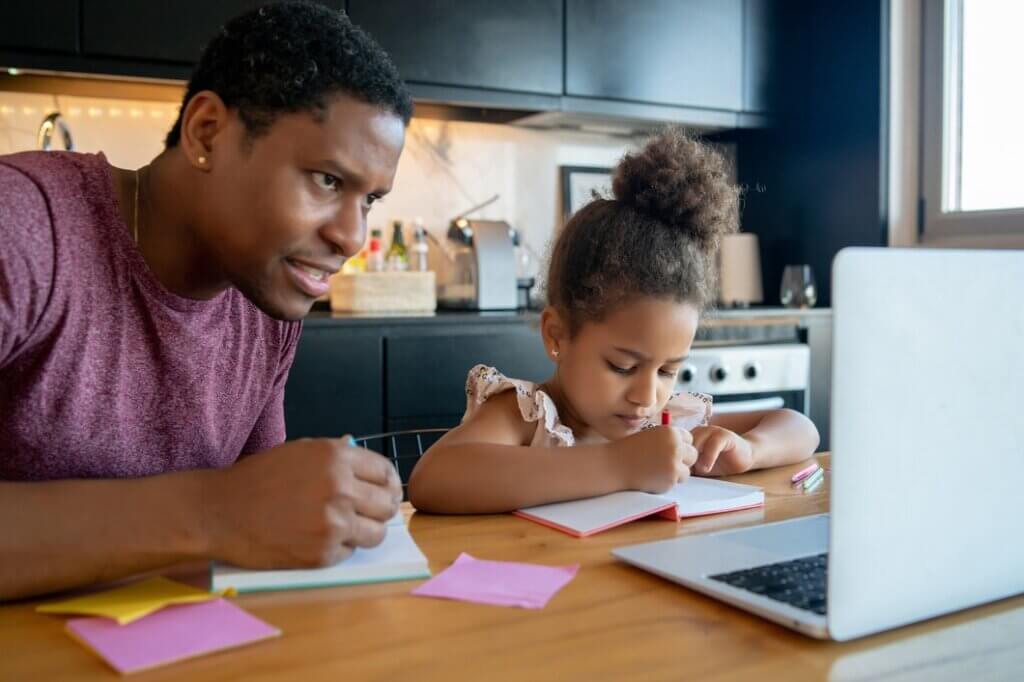 A parent and child engage in homework at a wooden table, surrounded by colorful sticky notes and a laptop. A modern kitchen is in the background.