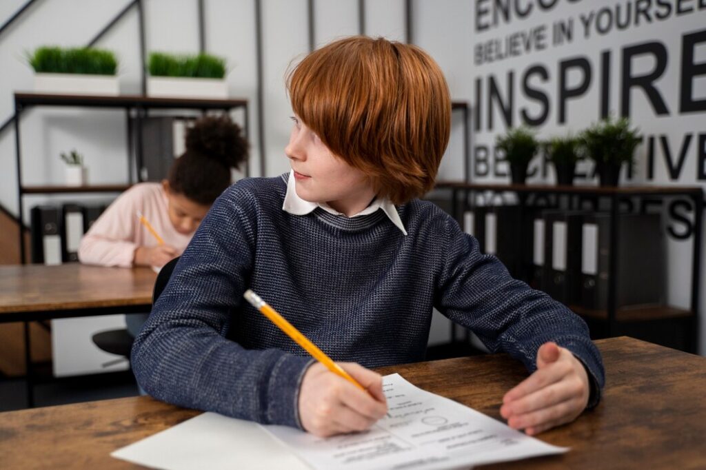 A red-haired boy in a blue sweater turns to look sideways while holding a pencil and sitting at a desk, seemingly pondering "What is 11 Plus Exam?". Another student is seated in the background, writing. The classroom has motivational words on the wall and shelves with plants and binders.