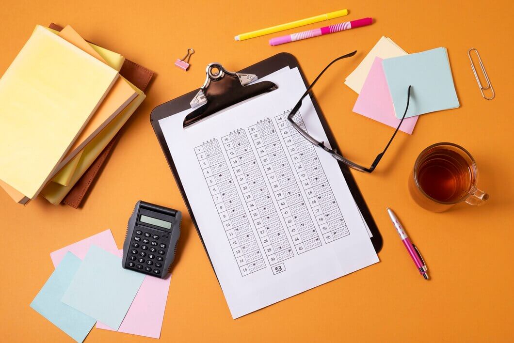 A clipboard with a printed calendar is surrounded by office supplies including a calculator, sticky notes, a stack of papers, pens, a pair of glasses, a paper clip, a binder clip, and a cup of tea, all arranged on an orange surface.