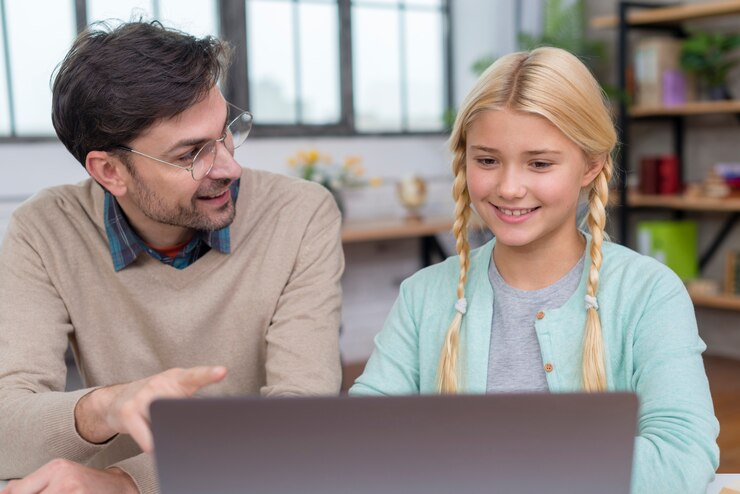 A man with glasses and a beard is sitting next to a young girl with blonde hair in braids. They are looking at a laptop screen together, appearing engaged and happy, showcasing the benefits of tutoring. The background shows a brightly lit room with shelves and windows.