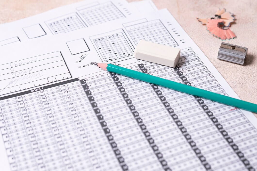 A standardized test answer sheet with filled and blank multiple-choice bubbles sits on a table. A green pencil, an eraser, pencil shavings, and a sharpener are on top of the sheet. Handwritten notes are partially visible in the background.