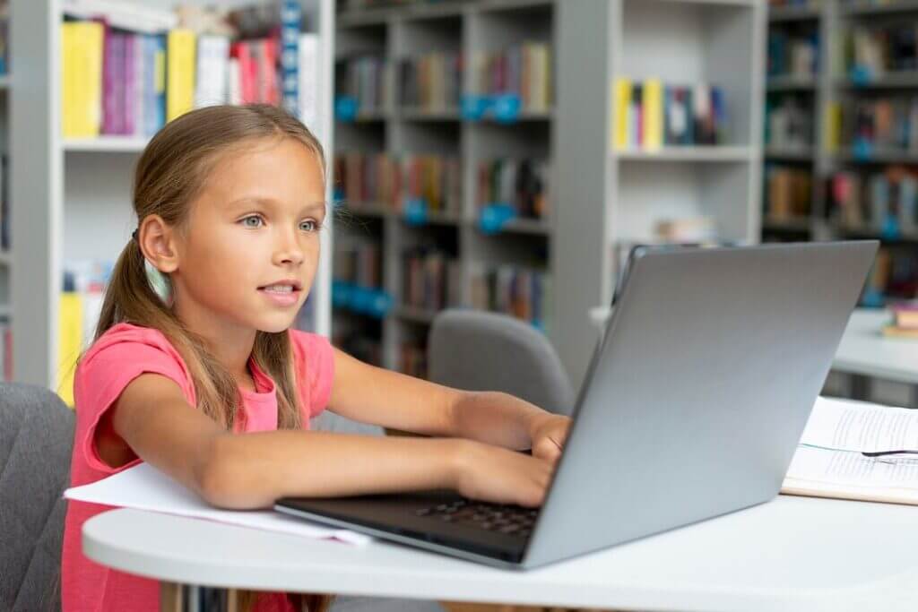 A young girl with long hair sits at a table in a library, focused on a laptop in front of her. She is wearing a pink shirt and has paper documents scattered nearby. Bookshelves filled with books are visible in the background.