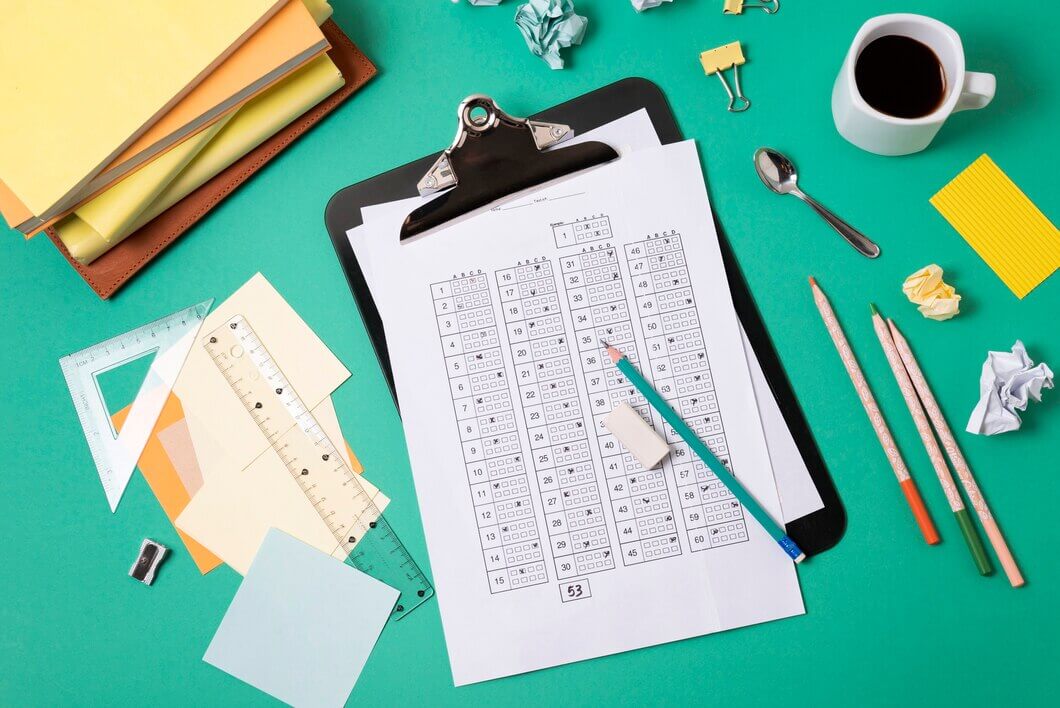 A cluttered desk with a green surface, featuring a clipboard holding a math worksheet filled with numbers, a pencil, rulers, crumpled paper, sticky notes, a cup of coffee, paper clips, and colorful pencils.