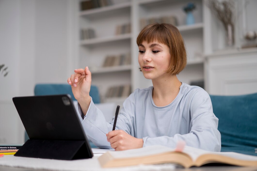 A person with short brown hair is sitting at a table with an open book and a tablet, using a stylus to interact with the screen. Shelves filled with books and decorative items are visible in the background. As they research, you might wonder, "How much does a science tutor cost?