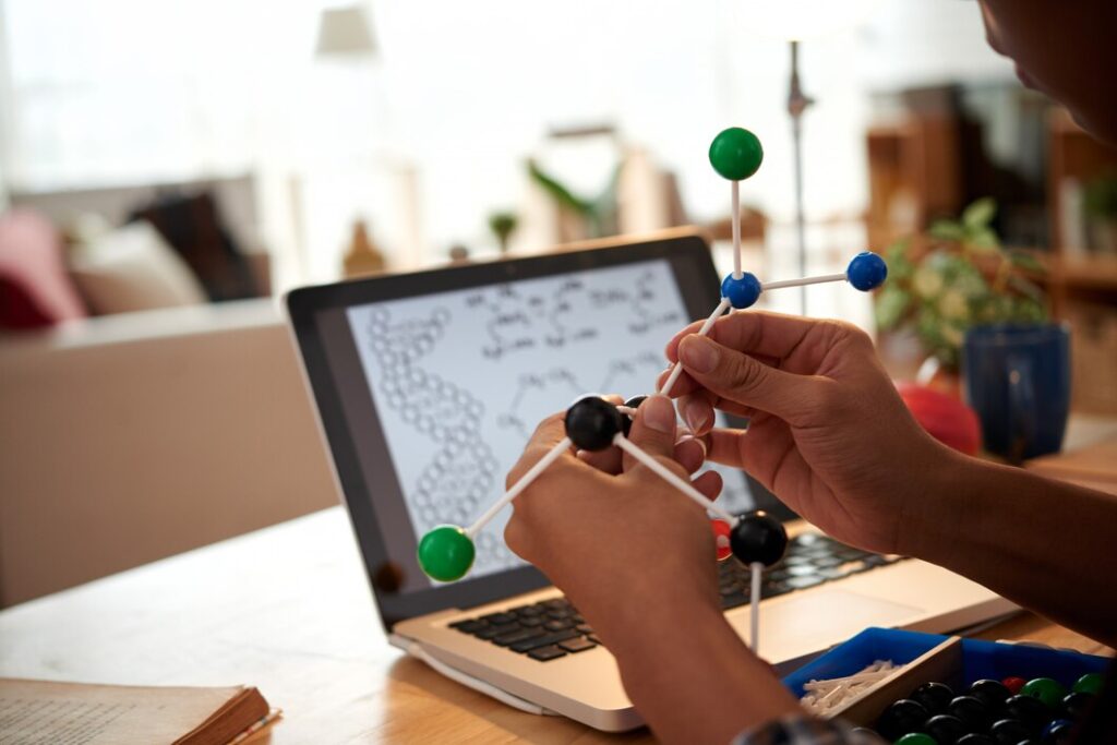 A person holds a molecular model while sitting at a desk with an open laptop displaying a science diagram. The background has blurred elements of a modern interior, including a plant and furniture, creating a focused workspace atmosphere—ideal for wondering how much does a science tutor cost.