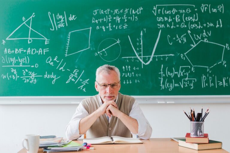 A senior man with white hair and glasses, dressed in a beige sweater vest over a white shirt, sits at a desk with an open book titled "How to Prepare for A Level Maths." Behind him, a green chalkboard is filled with complex mathematical formulas and geometric diagrams. Books and pencils are on the desk.