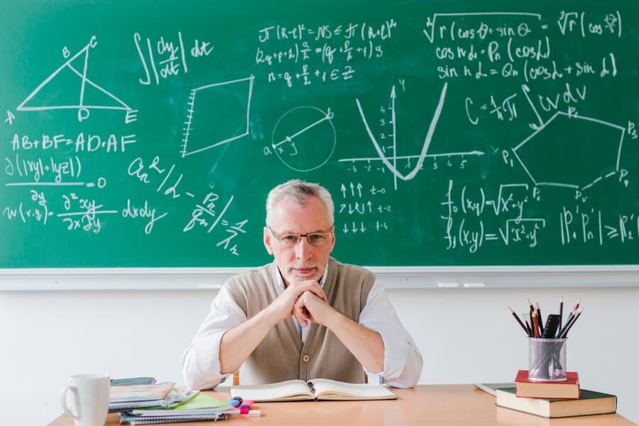 A senior man with white hair and glasses, dressed in a beige sweater vest over a white shirt, sits at a desk with an open book titled "How to Prepare for A Level Maths." Behind him, a green chalkboard is filled with complex mathematical formulas and geometric diagrams. Books and pencils are on the desk.