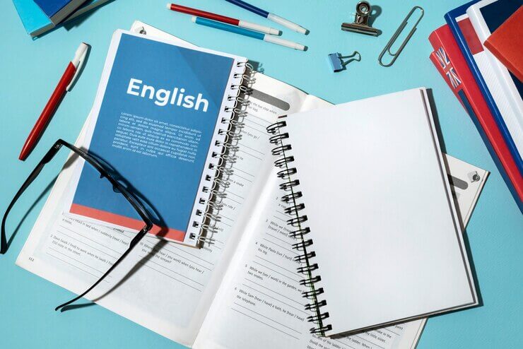 An English book, pen, glasses, and notebook arranged with office supplies on a vibrant blue background.