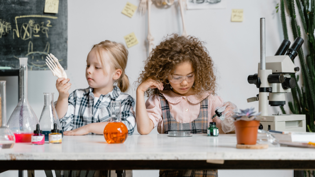 Two kinesthetic learners explore a science classroom. One holds a model skeleton hand, while the other examines a petri dish. Surrounded by lab equipment like beakers and a microscope, notes and a plant grace their table as hands-on discovery fills the air.
