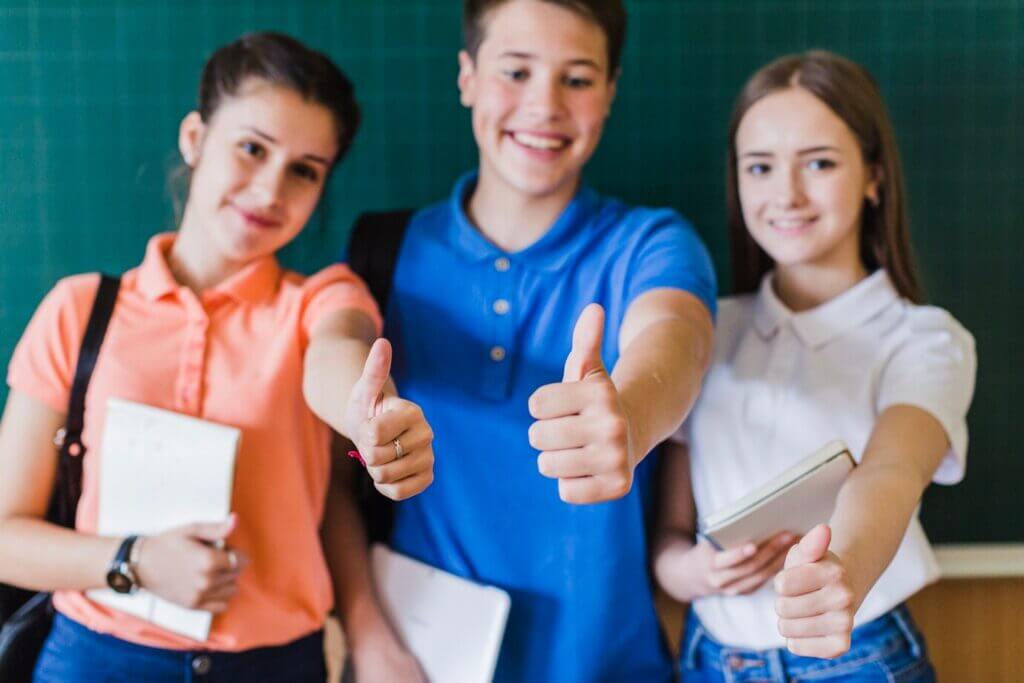 Three secondary school students smiling and giving thumbs up in front of a blackboard, showcasing enthusiasm and teamwork.