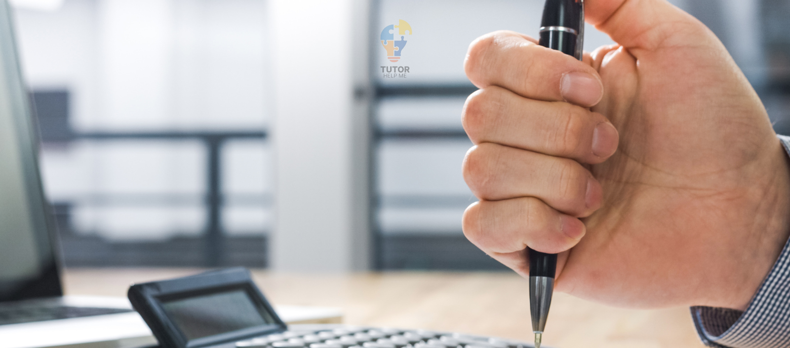 A close-up of a hand holding a pen, poised above a calculator on a wooden desk, with a laptop in the background.
