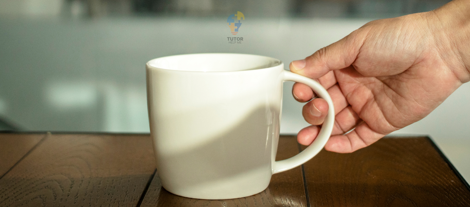 A hand reaches for a white coffee mug sitting on a wooden table, with a blurred background. The mug is empty and cylindrical.