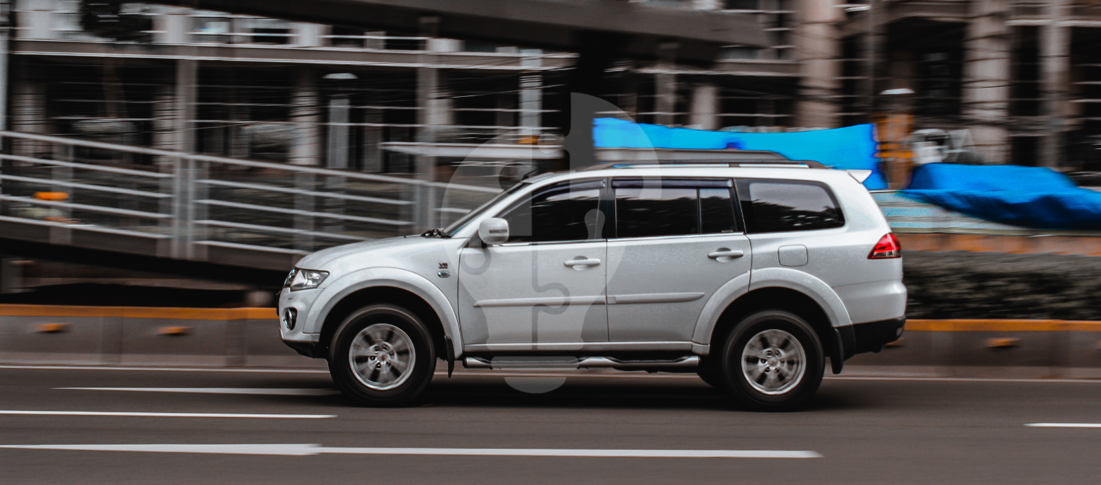 A silver SUV speeds past on a city street with blurred background, suggesting motion in an urban setting.