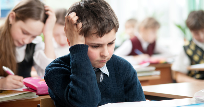 A young student sits at a desk in a classroom, resting their head on one hand, appearing thoughtful or stressed among classmates.