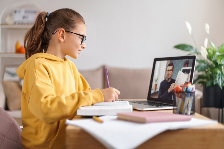 A student in a yellow hoodie takes notes during an online lesson on a laptop, surrounded by study materials on a wooden table.