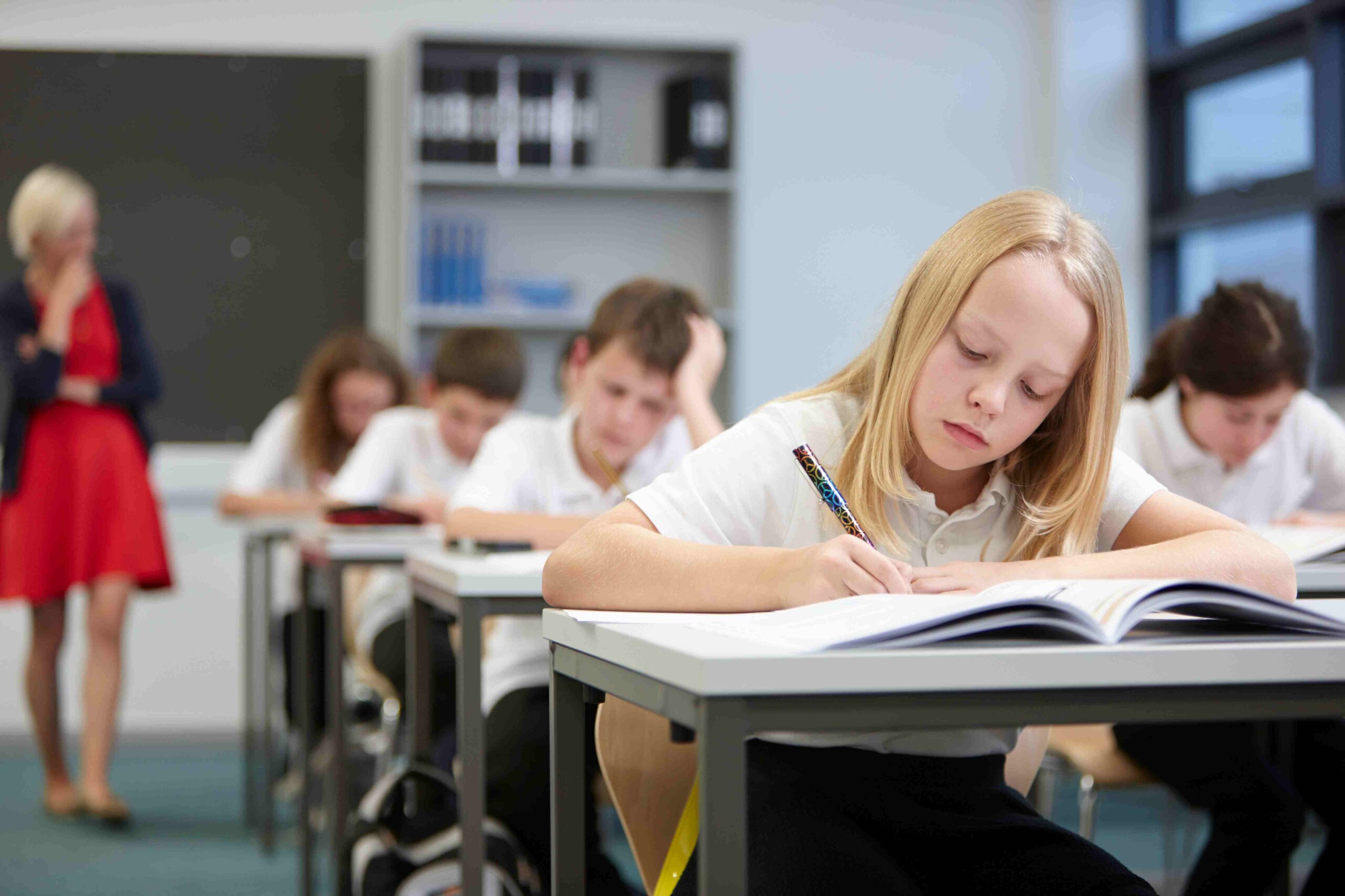 A classroom setting where students in white shirts are focused on their work, while a teacher observes in the background.