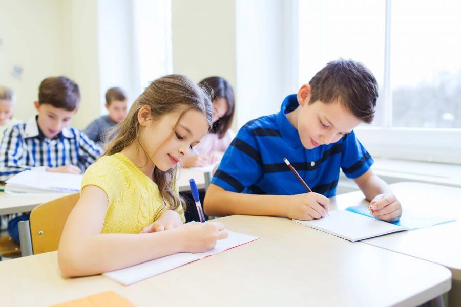 A group of children sitting at desks in a classroom, focused on writing on paper. They are of diverse backgrounds, wearing various clothing styles, and are engaged in their learning activities. The classroom is equipped with tables and desks against a wall.