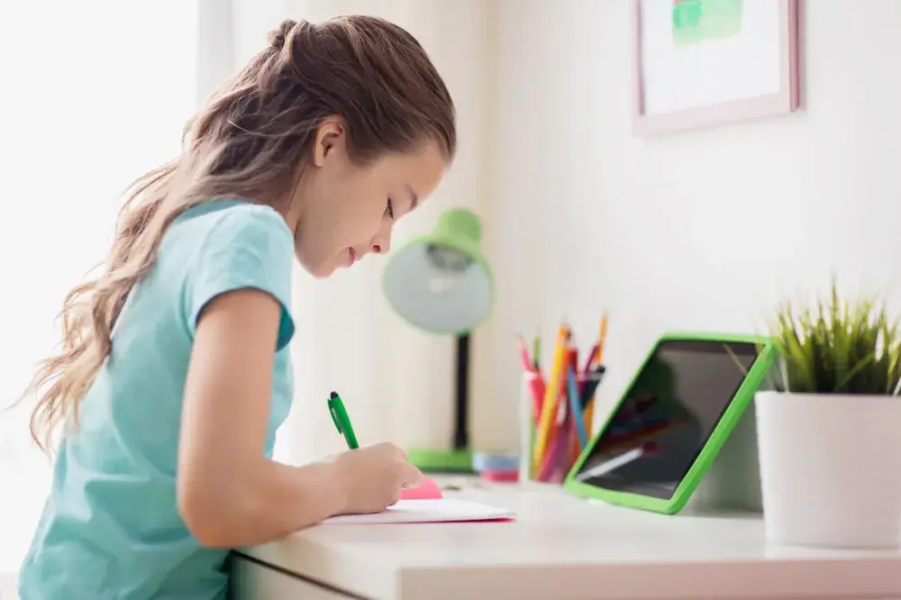 A child is sitting at a desk, writing on a notepad with a green pen, surrounded by colorful stationery and a tablet.