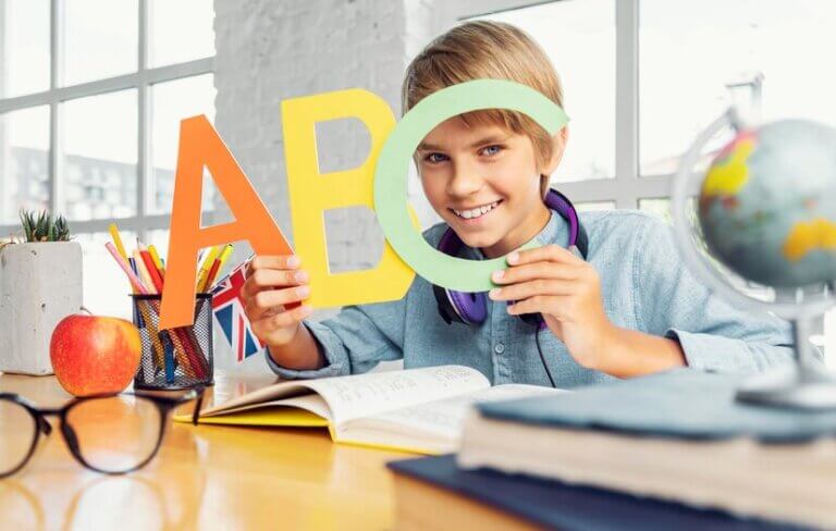 A child holds colorful letters 'A', 'B', and 'C' while studying at a desk with books, an apple, and a globe in a bright room.