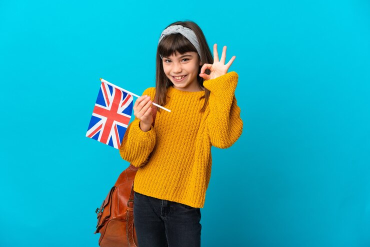 A child in a yellow sweater holds a small British flag and smiles, standing against a bright blue background with a brown backpack beside them.