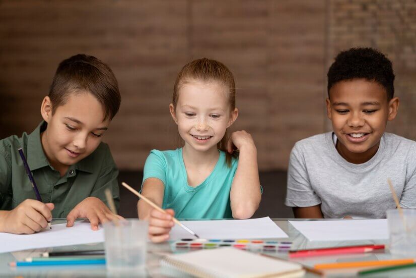 A group of children sitting at a table in a classroom, smiling and engaged in learning activities. They are wearing various clothing and include both girls and boys, with some focused on art.