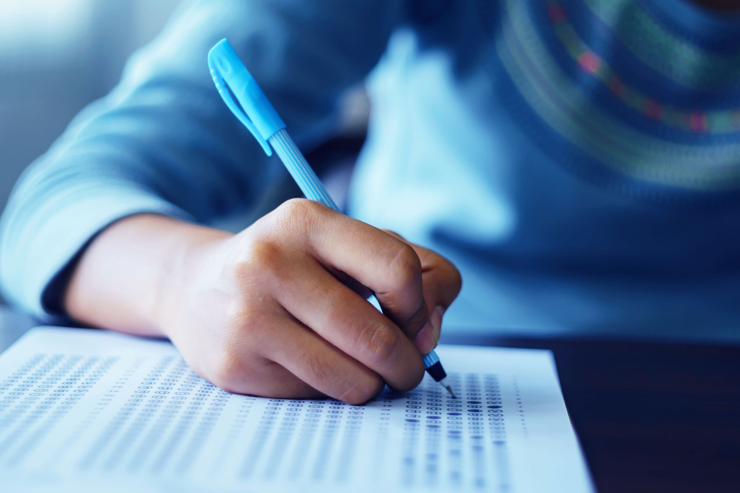A close-up of a hand holding a blue pen, filling out a printed answer sheet filled with bubbles and text.
