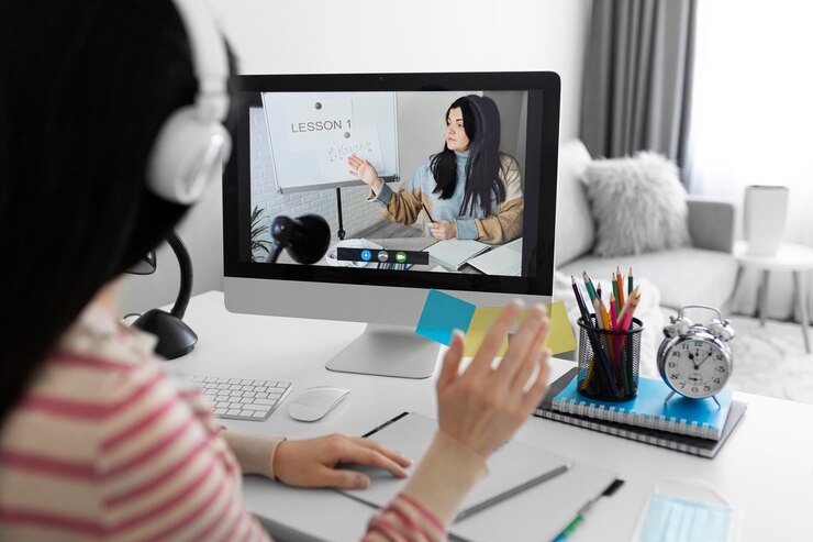 A person wearing headphones waves while participating in an online lesson displayed on their computer screen. A tidy workspace is visible.