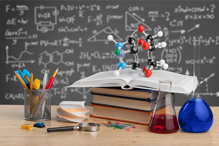 Desk scene with books, a molecular model, lab glassware, and stationery against a chalkboard filled with scientific equations.