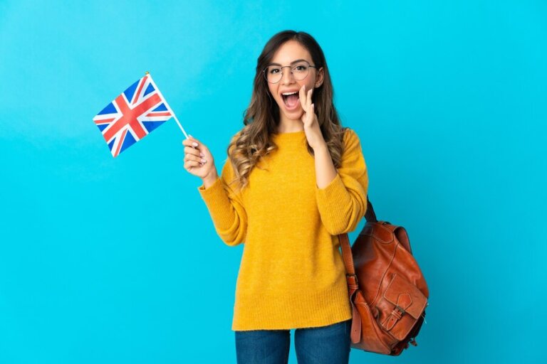A person wearing a bright yellow sweater holds a small British flag, standing against a light blue background, with a brown backpack at their side.