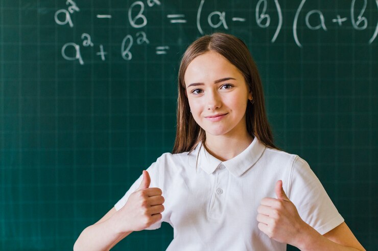 A student in a white polo shirt gives a thumbs up in front of a chalkboard filled with mathematical equations.