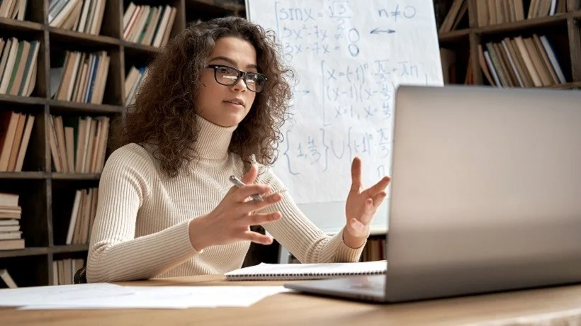 A person in a beige sweater gestures animatedly during an online meeting, with a laptop and notebooks on the table. Bookshelves are in the background.