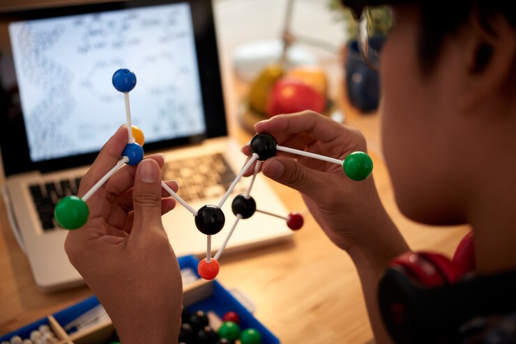 A person holds a colorful molecular model, with a laptop displaying scientific diagrams in the background.