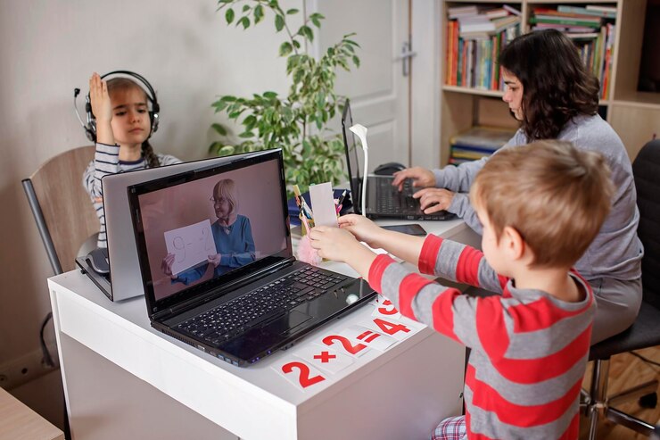 Children engage in online learning, with one boy showing a paper to a laptop while equations are displayed on the table.