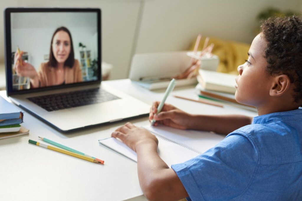 A child writes in a notebook while attending an online class, with a laptop displaying a teacher. Colorful pencils are nearby.