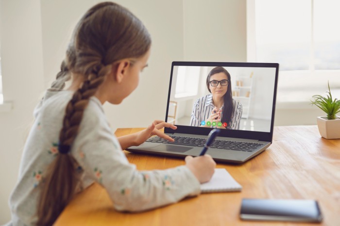 A young girl is engaged in an online class, taking notes while sitting at a wooden table with a laptop and a potted plant nearby.