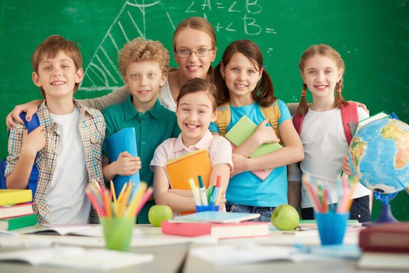 A group of schoolchildren stands together in a classroom, surrounded by books, stationery, and a globe, against a green chalkboard.