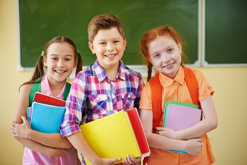 Three children stand in front of a chalkboard, holding colorful folders and books, smiling and dressed in casual school attire.