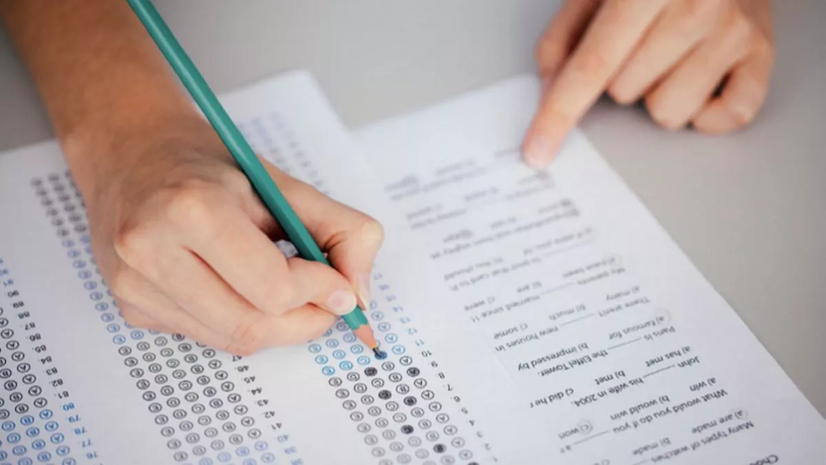A hand holding a pencil marks answers on a standardized test sheet with multiple-choice questions and answer bubbles.