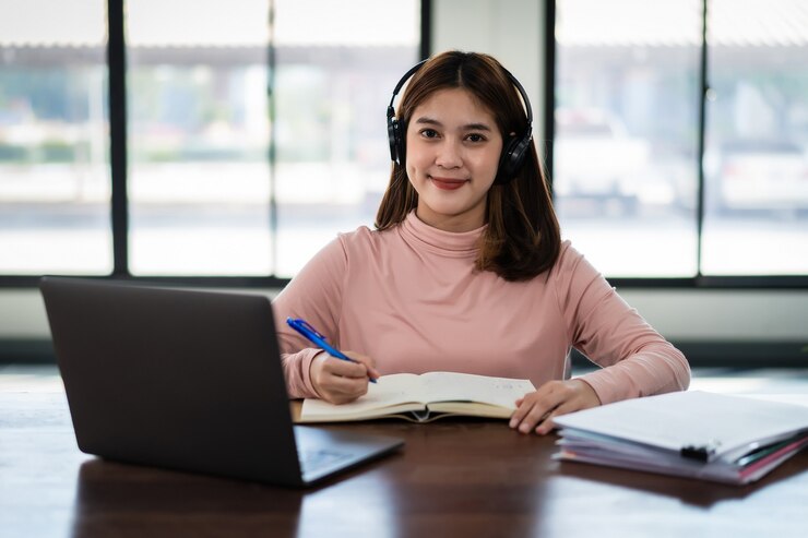 A person wearing headphones sits at a desk, writing in a notebook, with a laptop and documents in front of them, illuminated by natural light.