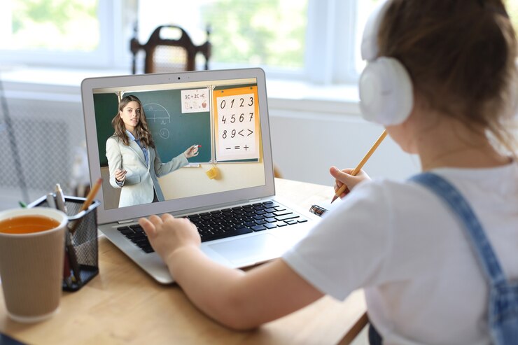 A child wearing headphones listens to an online math lesson on a laptop, taking notes with a pencil at a wooden desk.