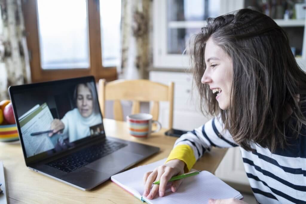 A person writing in a notebook while participating in an online video call, surrounded by a cup and fresh fruit on a table.