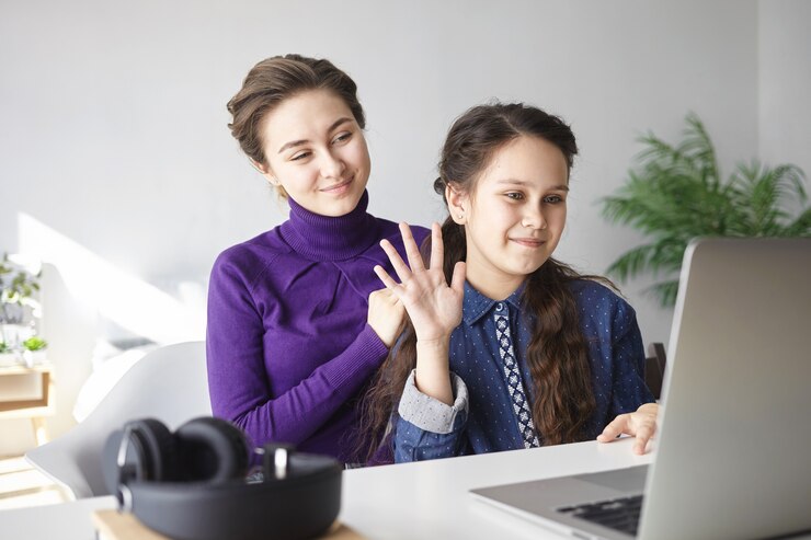 A woman and a girl interact at a computer, with a headset on the table, in a bright, modern workspace.
