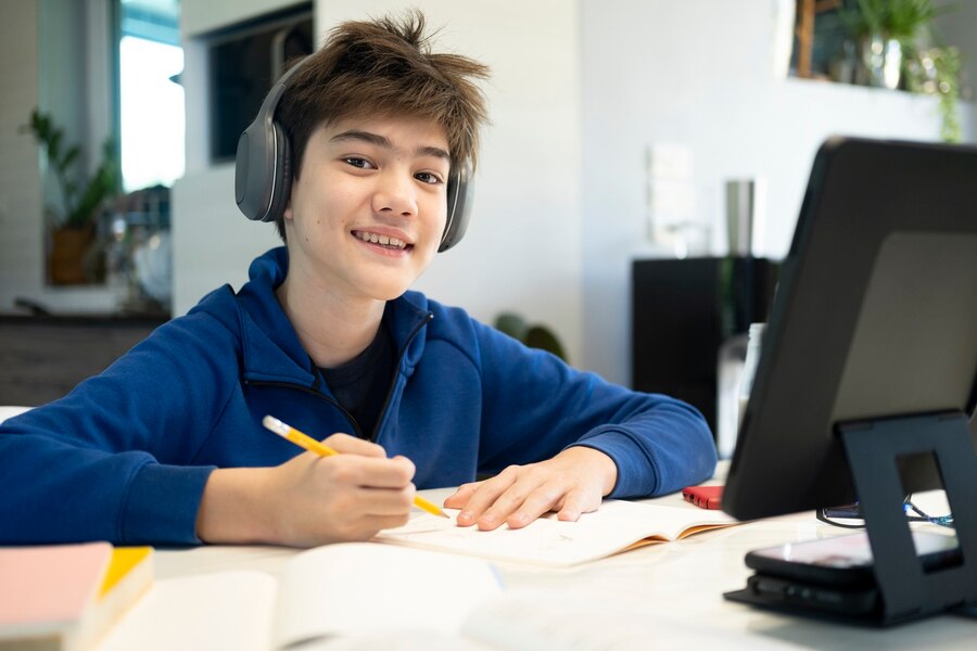 A student wearing headphones sits at a table, focused on writing notes in a notebook, with a digital tablet beside them.