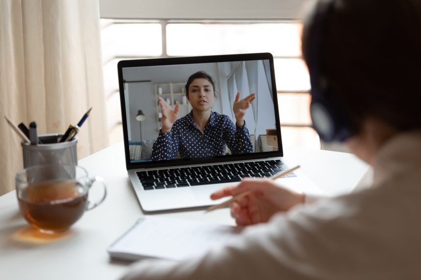 A person engages in a video call on a laptop, gesturing emphatically while the viewer takes notes, indicating an active discussion.