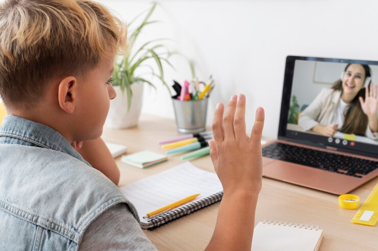 A child with curly hair sits at a desk, waving at a laptop during an online class, with school supplies and notebook nearby.