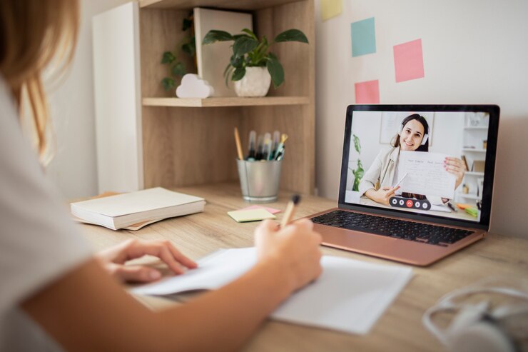 A person writing notes at a desk while participating in a video call on a laptop, surrounded by stationery and a plant.
